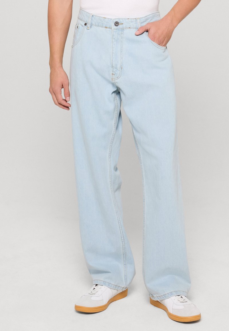 Vaaleansiniset farkut suoran leikkauksen omaavina, joissa on etutaskut, nappisuljenta ja vaaleat ompelun yksityiskohdat, yhdistettyinä valkoisiin tennareihin.