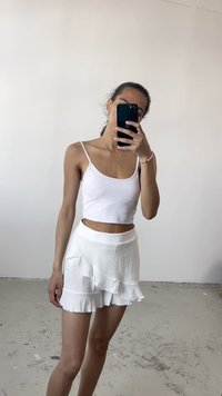 Weißes, kurzes Tanktop kombiniert mit einem weißen Minirock mit Rüschen. Der Rock hat einen strukturierten Stoff und schichtweise Rüschen am Saum.