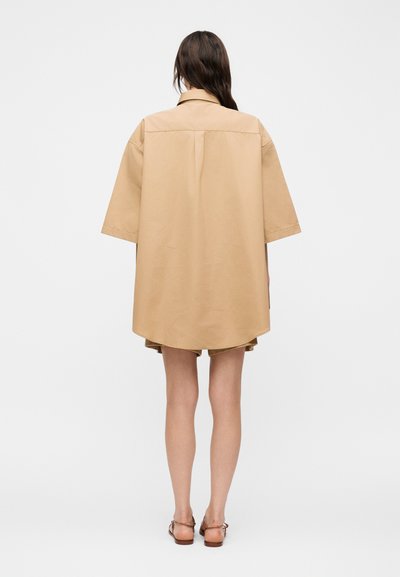 Γυναίκα στέκεται με την πλάτη στην κάμερα φορώντας υπερμεγέθη μπεζ πουκάμισο-φόρεμα και καφέ σανδάλια με λουριά σε απλό λευκό φόντο.