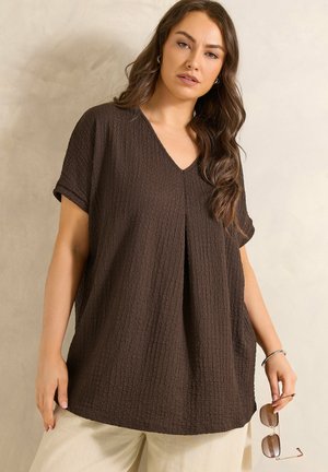 Kobieta z długimi falującymi włosami nosi luźną, ciemnobrązową, teksturowaną bluzkę z dekoltem w serek i beżowe spodnie, trzymając okulary przeciwsłoneczne w prawej ręce.