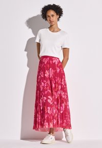 Fließender, plissierter Rock mit einem pinken Blumenmuster, kombiniert mit einem weißen T-Shirt und weißen Sneakern. Die Rocklänge reicht bis zur Mitte der Waden.