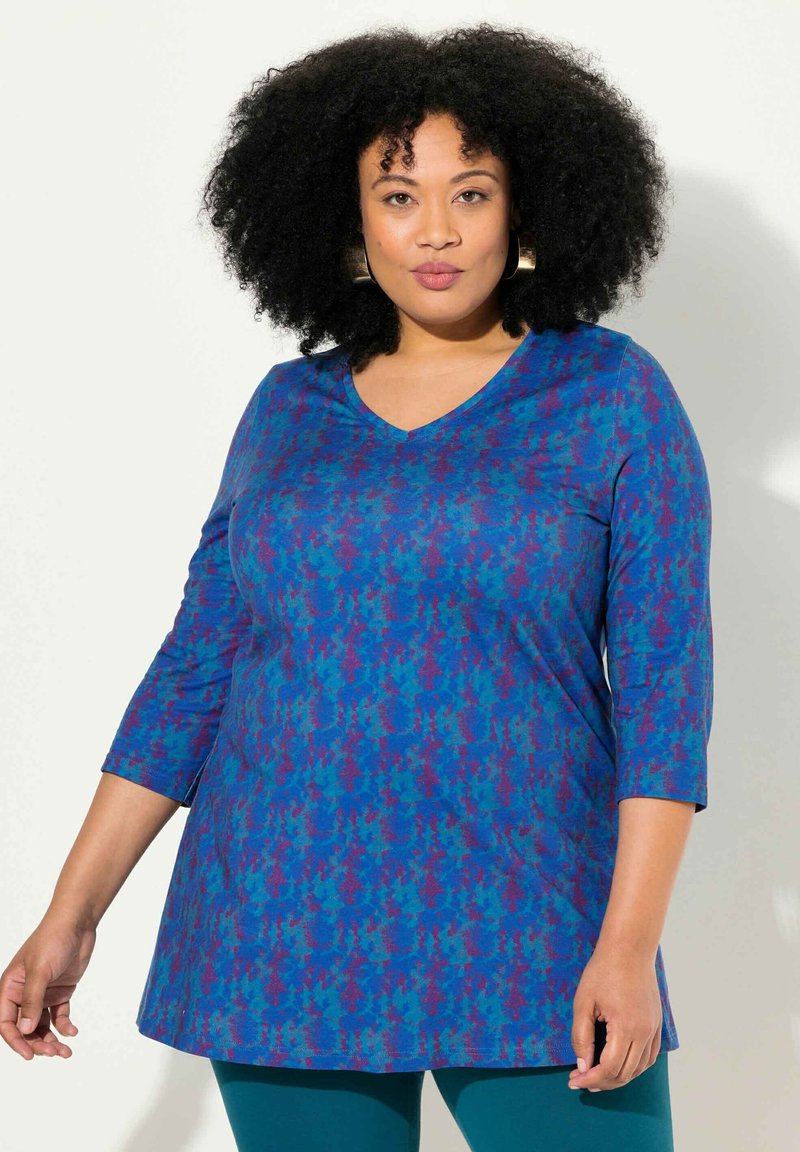 Sininen V-kauluksinen tuniikka, jossa on violetti houndstooth-kuvio. Valmistettu pehmeästä kankaasta, jossa on kolmannespituiset hihat ja rento muotoilu.