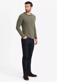 Olivgrüner, strukturierter Pullover mit langen Ärmeln, dunkle Jeans, braune Wildleder-Stiefeletten, taillierte Passform, Seitenansicht, Hände in den Taschen.