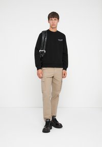 Black sweatshirt with ribbed cuffs, beige cargo pants with side pockets, and black chunky sneakers. Simple buckle shoulder strap adds detail.