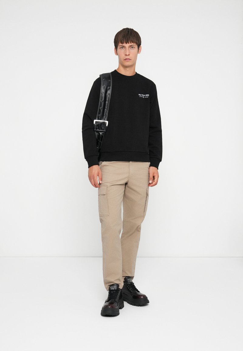Sweatshirt noir avec des poignets côtelés, pantalon cargo beige avec des poches latérales et baskets noires chunky. Une bandoulière simple à boucle ajoute du détail.