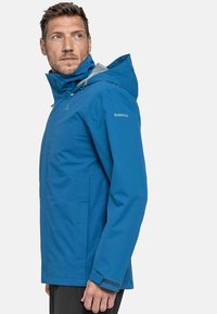 Chaqueta impermeable azul con un alto cuello, capucha ajustable y bolsillos con cremallera. Presenta acentos grises y un diseño de corte relajado.