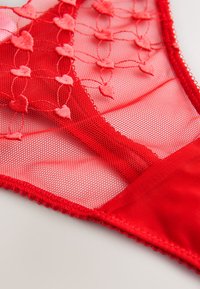 Rødt tyndt stof med hjerteformet broderi og bølgede kanter, detaljeret nærbillede af delikat lingerimateriale.