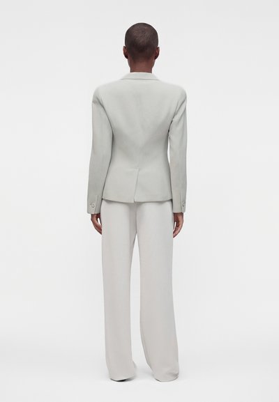 Personne debout de dos face à la caméra, portant un blazer gris clair ajusté et un pantalon large, sur fond blanc uni.