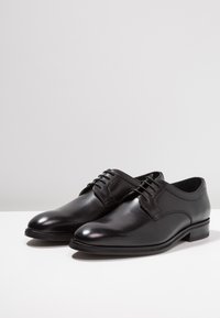 Zapatos de vestir de cuero negro con un acabado suave, punta redonda y sistema de cordones. Presentan un tacón bajo y sutilezas en la costura.