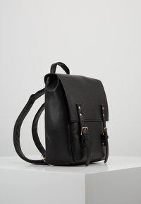 Zaino in pelle nera con patta pieghevole, dettagli in metallo dorato, spallacci regolabili e due fibbie frontali. Texture liscia, forma strutturata.