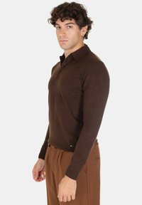 Polo shirt marrone a maniche lunghe con colletto, polsini a costine e una leggera texture. La vestibilità è sagomata, indossato con pantaloni marroni chiari.
