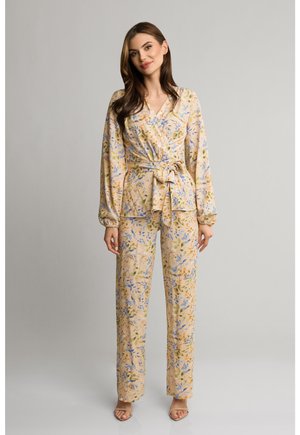 Kobieta stojąca, ubrana w beżową kwiecistą kopertową bluzkę z długimi rękawami i dopasowane szerokie spodnie, pozująca na jednolitym tle.