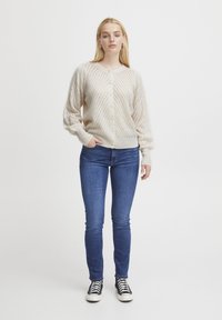 Cardigan en tricot beige avec un motif en chevron, présentant un col rond, des manches longues et une fermeture à boutons. Porté avec un jean bleu et des baskets noires.