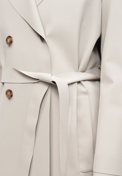 Ανοιχτό μπεζ παλτό διπλού στήθους με κουμπιά σε σχέδιο χελωνόσκεπας, δεμένη υφασμάτινη ζώνη και πλαϊνή τσέπη στα δεξιά.