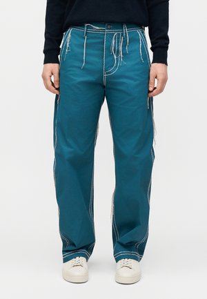 Osoba nosząca turkusowe spodnie o szerokich nogawkach z białym kontrastowym przeszyciem i postrzępionymi nitkami, w połączeniu z białymi trampkami i czarnym swetrem.