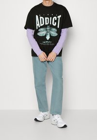 Černé grafické tričko s nápisem "NIGHT ADDICT" a designem můry, přes které je vrstveno levandulové tričko s dlouhým rukávem. Doplněno tyrkysovými kalhotami a bílými teniskami.