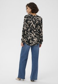 Blouse noire avec un motif abstrait beige, manches longues et détail froncé à l'arrière, associée à un jean large bleu clair.