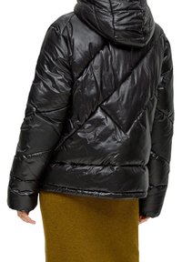 Sort pufferjakke med en blank overflade, diagonal quiltet mønster og hætte. Har ribbede manchetter og en tætsiddende kant, båret over en grøn strikket nederdel.