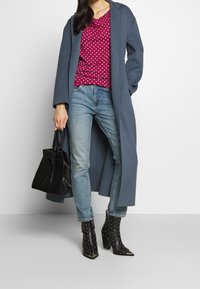 Lång grå kappa, röd mönstrad topp, blå jeans, svarta nitar ankelskor, bärande en svart väska. Har en avslappnad passform och olika texturer.