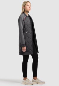 Žena stojí a má na sobě šedou prošívanou dlouhou bundu, černé legíny a světle béžové tenisky na jednobarevném šedém pozadí.