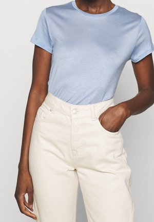 Mujer llevando una camiseta de manga corta azul claro metida en pantalones de talle alto de color crema, con una mano en el bolsillo.