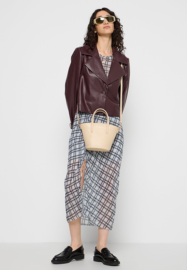 Frau mit großen Sonnenbrillen, bordeauxroter Lederjacke, kariertem Kleid mit Schlitz, schwarzen Mokassins und beiger Handtasche mit Schultergurt.