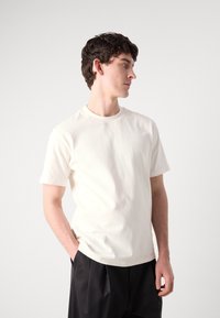 Short-sleeve, crew-neck t-shirt in off-white cotton. Features minimal branding in the center, with a relaxed fit and smooth texture.