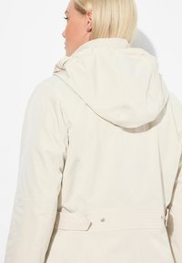 Beige mikina s kapucí vyrobená z hladkého, lehkého materiálu; obsahuje jemné švy a stažený pas se zapínáním na patentku.
