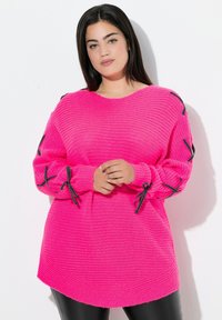 Eine Frau trägt einen leuchtend pinkfarbenen Strickpullover mit schwarzen Satin Schleifen an den Ärmeln und steht vor einem weißen Hintergrund.