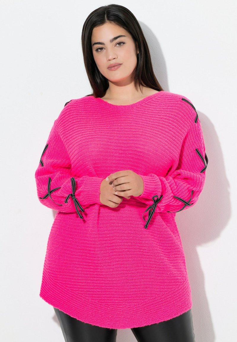 Eine Frau trägt einen leuchtend pinkfarbenen Strickpullover mit schwarzen Satin Schleifen an den Ärmeln und steht vor einem weißen Hintergrund.