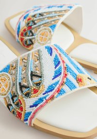 Sandales à glissière blanches avec un travail de perles multicolores complexe présentant des motifs géométriques et abstraits sur les larges brides avant.