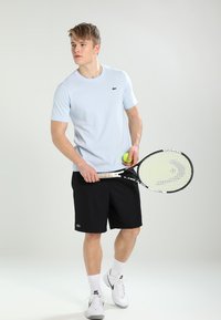Világoskék, rövid ujjú sportpóló zöld logóval, fekete sort, teniszütőt és labdát tart, fehér sportcipő, sportos szabás.
