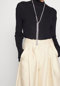 Top nero a maniche lunghe abbinato a pantaloni a gamba larga color crema; un collana di catena con strass argento adornano il collo. Tessuti lisci e vestibilità su misura.