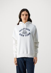 Felpa bianca con un stampa frontale che recita "HILFIGER est 1985 NEW YORK" in blu navy. Presenta un cappuccio con cordino e polsini a coste. Realizzata in morbido misto cotone.
