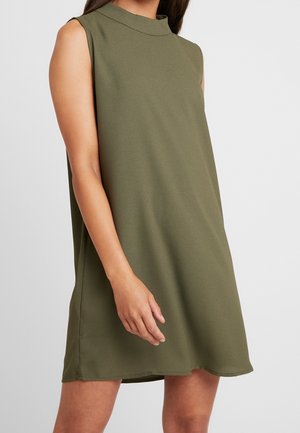 Femme portant une robe ample sans manches, col montant, couleur vert olive, debout avec une main près de sa cuisse sur un fond clair uni.