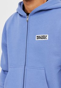 Niebieska bluza z kapturem na zamek, wykonana z miękkiego materiału, z przednią kieszenią, kapturem ze sznurkiem oraz czarno-białym logo "MULTIPLY APPAREL".