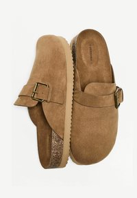 Rjavi semiš natikači s ploskim podplatom, z zaobljenim prstom in bočnim trakom z zaponko. Teksturiran detajl iz plute ob robu podplata.