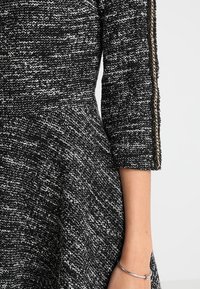 Robe texturée en noir et blanc avec des manches trois-quarts ornées d'un détail chaîne dorée le long du bras, portée avec un bracelet argenté au poignet.