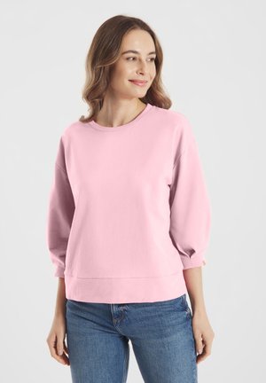 Vrouw met lichtbruin haar, gekleed in een loszittende roze top met lange mouwen en een blauwe spijkerbroek, kijkt iets opzij met een lichte glimlach.