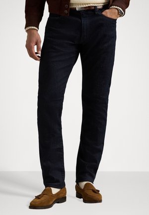 Mann trägt dunkle Jeans, braune Wildleder-Tassel-Loafer, beige Socken und einen braunen Pullover mit sichtbarem beigen Ärmelbund.