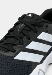 Sort atletiksko med mesh-øvre, der har tre hvide striber og teksturerede detaljer. Inkluderer en logoetiket og sorte snørebånd.