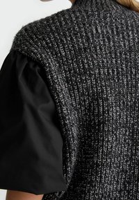 Tlustá pletená vesta v černé a šedé barvě, s texturovanou látkou a vysokým límcem, doplněná nadýchaným černým topem s rukávy pro vrstvený vzhled.