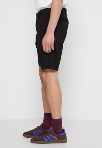 Homme portant un short noir, des baskets Adidas rouge foncé avec des bandes bleues, et des chaussettes assorties rouge foncé, debout sur un fond clair uni.