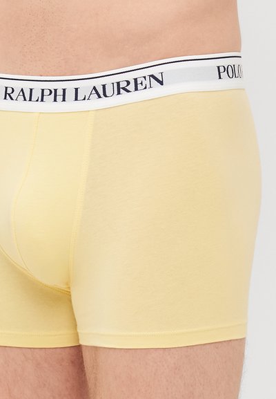 Rumene bombažne bokserice z belim in mornarsko modrim pasom, ki prikazuje blagovni znamki "Ralph Lauren" in "Polo". Gladka tekstura, oprijet dizajn.