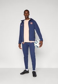 Blå sportsjakke og bukser med pink accenter, tætsiddende design og Barca-logo. Holder en hvid og blå fodbold, iført sorte fodboldstøvler.