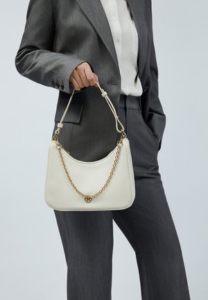 Witte leren handtas met een gebogen vorm, voorzien van een gouden kettingriem en een decoratieve hanger. De achtergrond is neutraal en minimalistisch.