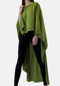 Grön, flödande cape med lös passform och asymmetrisk fåll. Tillverkad av slät tyg, bärs över svarta leggings, med mjuka veck och utan synliga stängningar.