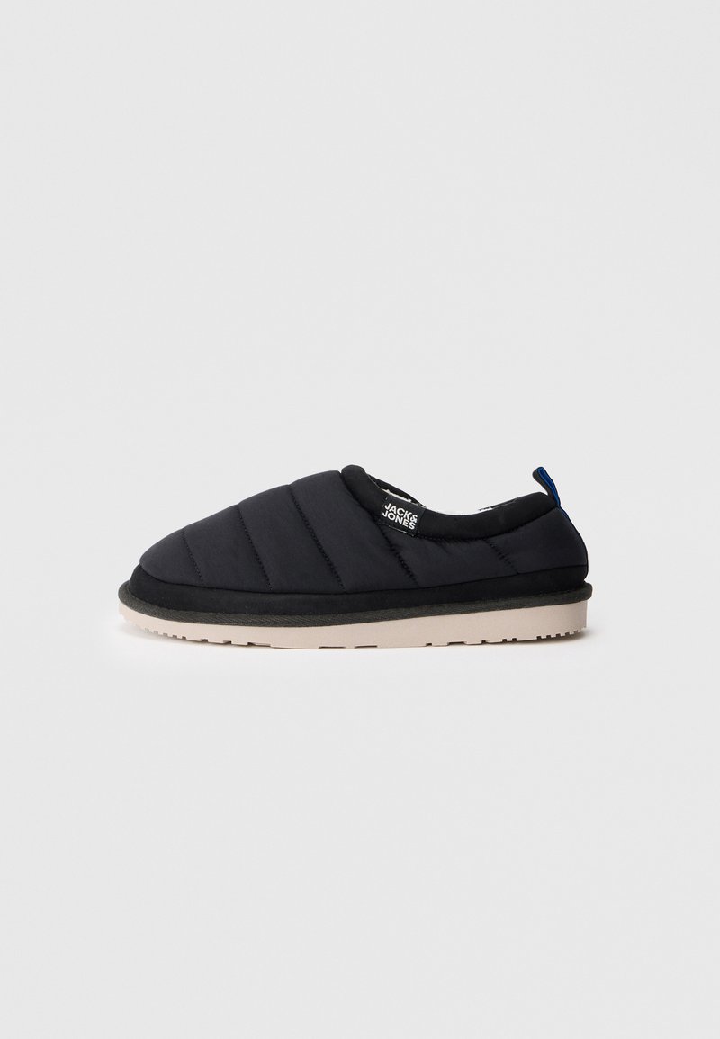 Schwarze Slipper mit einem gequilteten Obermaterial, einer abgerundeten Spitze, einem weichen Kragen und einer kontrastierenden weißen Sohle. Mit einem kleinen blauen Label an der Ferse.