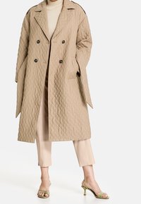 Abrigo beige acolchado de doble botonadura con grandes botones, bolsillos laterales y cuello ancho. Combinado con pantalones beige claros y sandalias verdes de tacón.