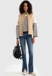 Beige prošívaná vesta přes bílé a námořnické pruhované tričko, spárovaná s rozšířenými modrými džínami a béžovými teniskami; drží černý batoh se zlatými akcenty.